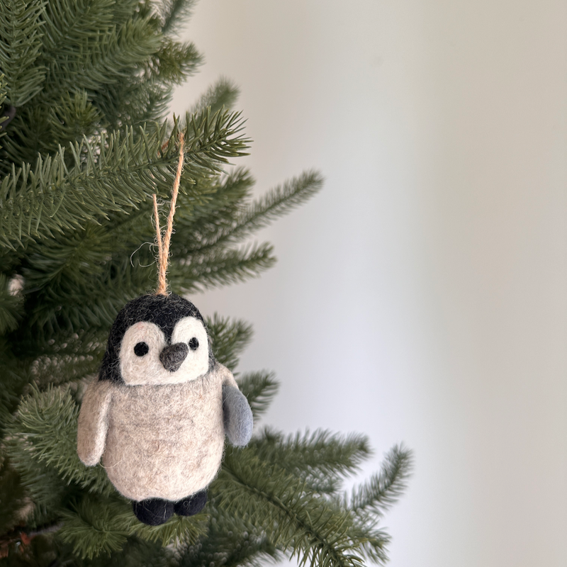 A charming needle felted penguin felt ornament is playfully hanging from a green Christmas tree branch. Set against a soft white background, this handcrafted felt ornament features a black head, beige body, and black feet, with one gray wing. A jute string is attached for easy display.