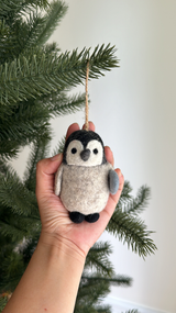A handmade felt penguin ornament is gently held by a female hand against the backdrop of a green Christmas tree. The playful, needle felted wool felt penguin features a grey body, black head, small black feet, and a gray wing, with black eyes and a small fish. A jute string loop at the top suggests it's ready to be hung as a unique Christmas tree decoration.