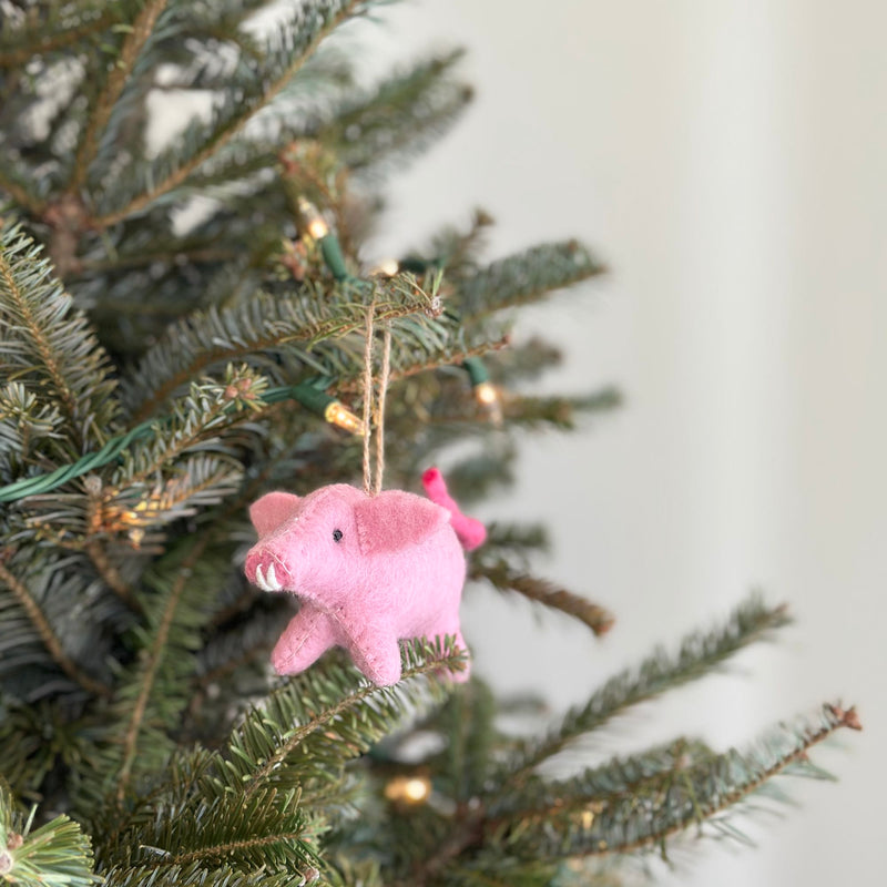 A charming needle-felted pig Christmas ornament hangs prominently on a Christmas tree, illuminated by small, warm white lights. This cheerful pink pig felt ornament is crafted from organic wool, featuring black eyes, a curly tail, and visible white tusks. A jute string is attached for easy display.