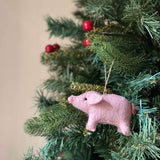 A pink needle-felted pig Christmas ornament hangs from a Christmas tree. Its rounded body, small ears, tiny black eyes, snout, and curly tail are visible among green needles and red berries. Crafted from organic wool, a jute string loop allows it to hang, adding a delightful, handmade touch to the festive decor.