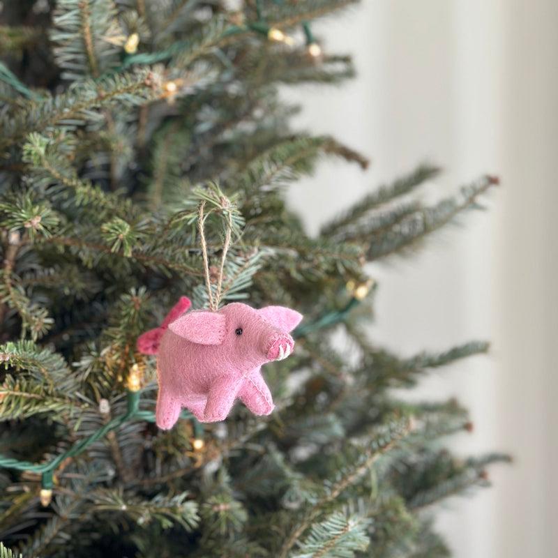 A light pink needle-felted pig ornament hangs festively on a Christmas tree. This soft, organic wool pig features a curly tail, pointy ears, and a snout, suspended by a jute string. Green Christmas tree branches with small light bulbs form the cozy backdrop.