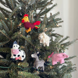 A handmade pink pig ornament hangs from a jute string on a brightly lit Christmas tree. This needle felted pig ornament, crafted from organic wool, features a small tail, floppy ears, and tiny facial details, adding a playful and festive touch to the tree.