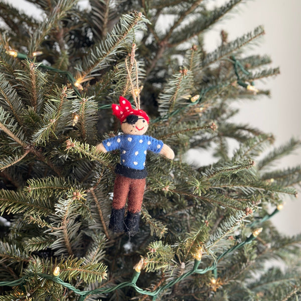 A felt pirate ornament hangs on a Christmas tree adorned with green needles and warm white lights. The rounded pirate ornament wears a blue shirt with white polka dots, brown pants, and a red bandana with white polka dots on its head. A jute string is attached for hanging, adding a festive and joyful touch to the tree.