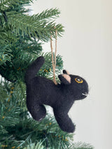 A playful wool felt cat ornament hangs from a vibrant green Christmas tree branch, suspended by a jute string. The black cat ornament features small pink inner ears, yellow outlined eyes, and white whiskers. It is presented against a soft white background, evoking a cozy and festive mood.