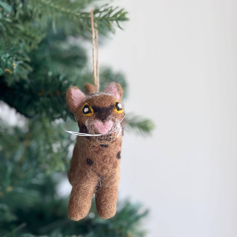 A playful cat wool felt ornament hangs from a green Christmas tree branch. This charming light brown cat ornament features small dark brown spots, light pink inner ears, a pink nose, and white whiskers, with a jute string for hanging.