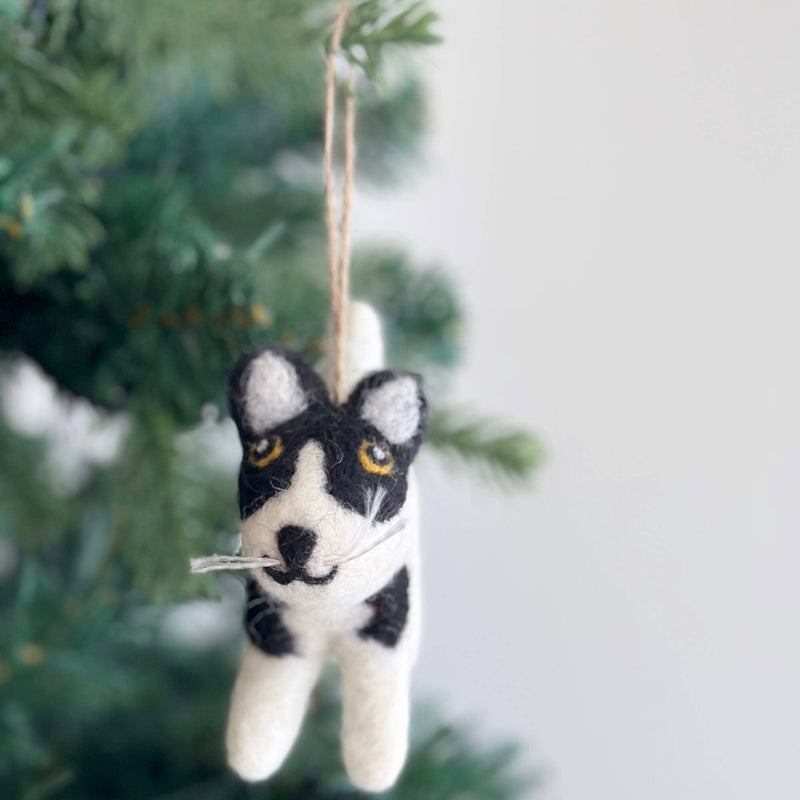 A playful wool felt cat ornament hangs from a Christmas tree. This handcrafted cat features black and white markings, a black smile, white whiskers, and yellow eyes. It is suspended by a jute string, with the cat playfully depicted hanging with its front paws up, adding a whimsical touch to holiday decor.