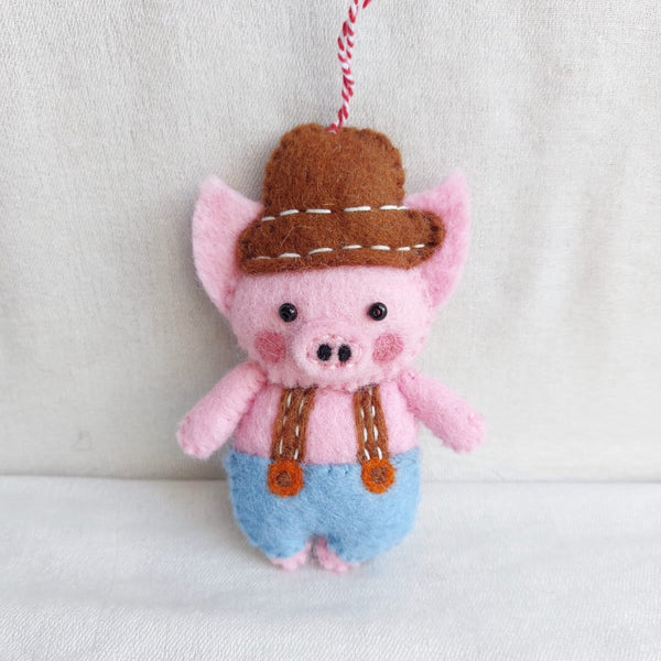 A wool felt plumber pig ornament rests against a soft white backdrop, conveying a playful and joyful mood. This handmade pink pig ornament features rosy cheeks, black eyes, and a brown hat with white stitching. It wears brown straps over blue overalls, fastened by orange buttons, and includes a red and white cotton string for hanging.