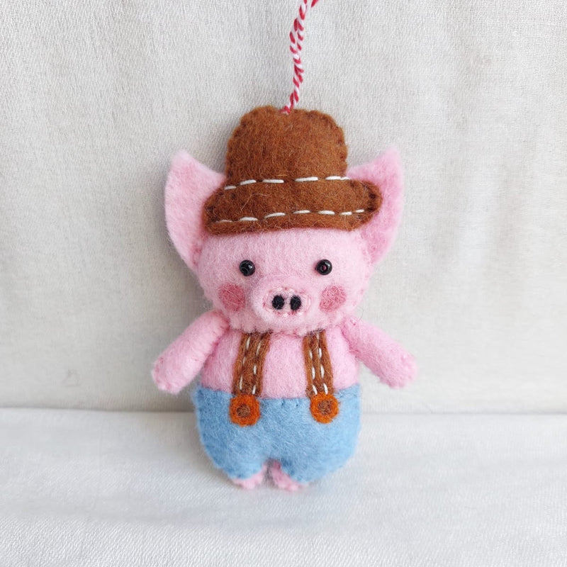 A wool felt plumber pig ornament rests against a soft white backdrop, conveying a playful and joyful mood. This handmade pink pig ornament features rosy cheeks, black eyes, and a brown hat with white stitching. It wears brown straps over blue overalls, fastened by orange buttons, and includes a red and white cotton string for hanging.