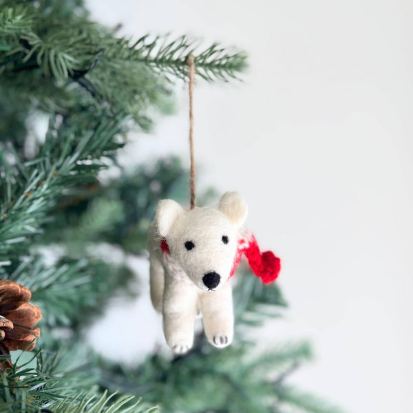 A fluffy white felt polar bear ornament hangs on a Christmas tree. This charming polar bear ornament wears a vibrant red scarf, with black bead eyes and nose. A jute string is attached for easy hanging.