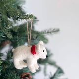 A white felt polar bear ornament, adorned with a red scarf, hangs from a green Christmas tree branch. This unique felt ornament is suspended by a jute string, set against the branch's green needles and pine cones, creating a festive ambiance.