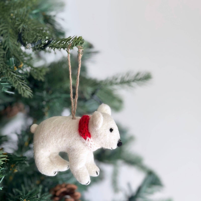 A white felt polar bear ornament, adorned with a red scarf, hangs from a green Christmas tree branch. This unique felt ornament is suspended by a jute string, set against the branch's green needles and pine cones, creating a festive ambiance.