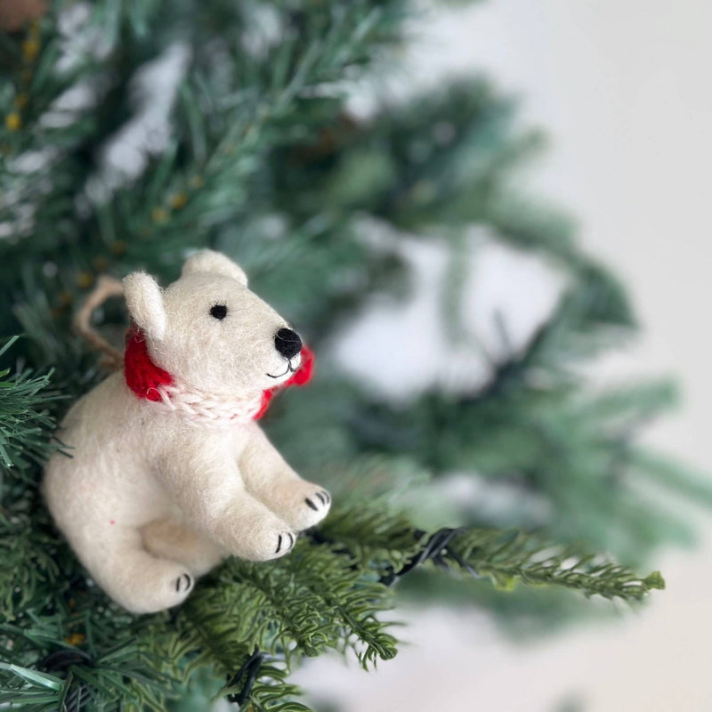 A felt polar bear ornament is perched on a vibrant green pine branch of a lush Christmas tree. This charming polar bear ornament has a rounded white body, black nose, and black feet. A festive red and white braided cotton string, resembling a scarf, is tied around its neck, and a jute string is attached for hanging.