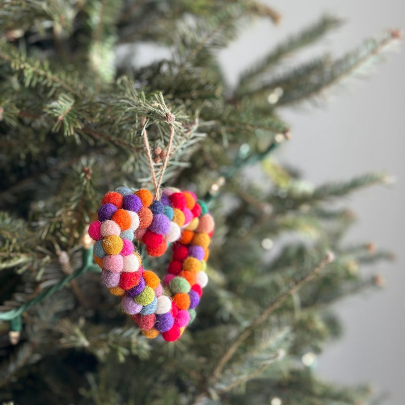 A colorful rainbow wool felt sheep ornament, crafted from numerous felt balls, hangs from a vibrant green Christmas tree. This unique needle-felted and hand-stitched sheep ornament showcases a cheerful, textured design in bright rainbow colors. A jute string allows this adorable sheep ornament to hang gracefully, adding a festive touch to holiday decor.