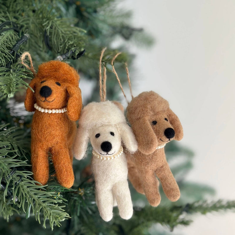 Three charming wool felt poodle ornaments, in white, cream, and brown, are displayed hanging on a green Christmas tree. Each handmade poodle ornament features a black nose, dark eyes, and a delicate pearl necklace, with a jute string attached for hanging.