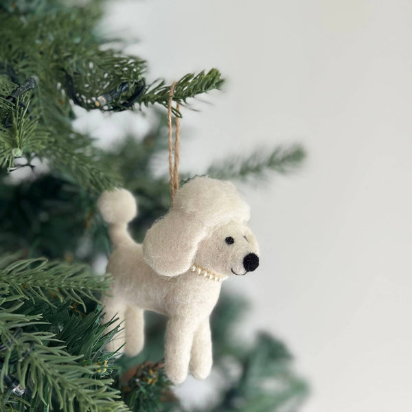 A white poodle wool felt ornament hangs elegantly from a lush green Christmas tree branch. This charming handcrafted felt ornament features a fluffy body, small black eyes, a black nose, and a sophisticated pearl necklace, suspended by a jute string.