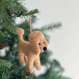 A light brown wool felt poodle ornament hangs from a Christmas tree branch. This charming poodle ornament features a rounded head with floppy ears, a small black nose, and is adorned with a pearl necklace. A jute string is attached to its head for hanging.