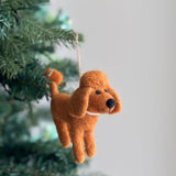 A brown handcrafted wool felt poodle ornament hangs from a Christmas tree branch, set against a softly blurred background. This charming felt poodle ornament features a textured, wool-like appearance, a round black nose, and a delicate pearl necklace around its neck.