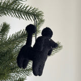 A wool felt black poodle ornament hangs from a Christmas tree branch. The felt poodle features a textured surface with a round ball on its head, topped by a smaller ball, and has four limbs, a tail, and a snout. A cotton string is attached for hanging.