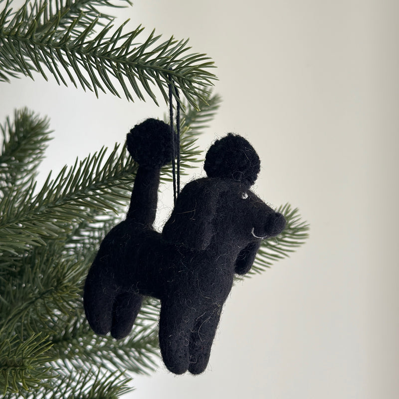 A wool felt black poodle ornament hangs from a Christmas tree branch. The felt poodle features a textured surface with a round ball on its head, topped by a smaller ball, and has four limbs, a tail, and a snout. A cotton string is attached for hanging.