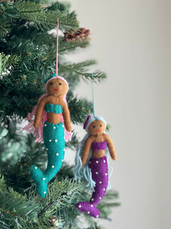 Two vibrant handcrafted felt Pride Mermaid ornaments hang gracefully on a Christmas tree with vivid green needles. One mermaid features light brown skin, pink hair, a turquoise top, and a matching tail adorned with white dots, suspended by a pink string. The second mermaid has light brown skin, light blue hair, a purple top, and a coordinating tail with white dots, hanging from a light blue string. These unique Christmas ornaments embody self-expression and radiant joy.