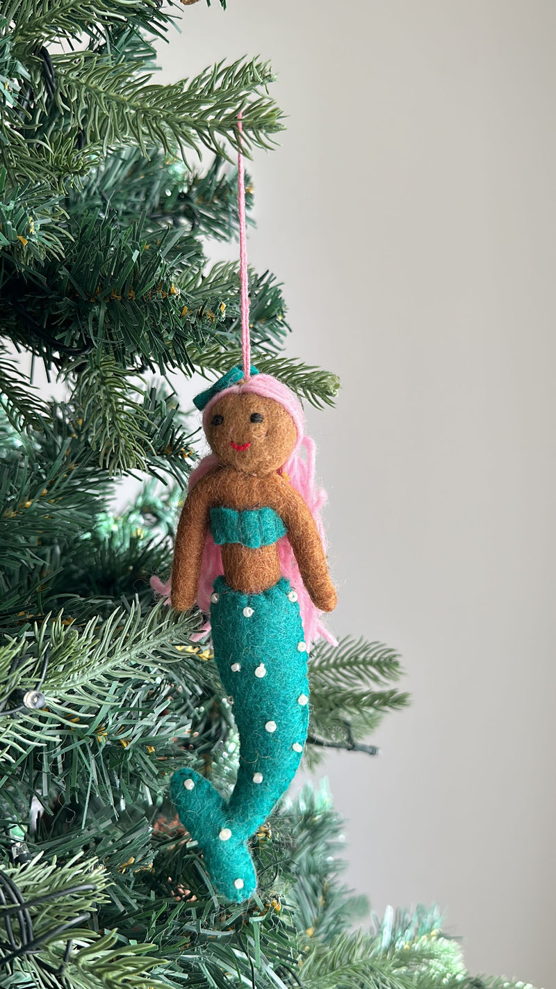 A handcrafted felt mermaid ornament hangs on a Christmas tree, set against a light gray background. This festive felt mermaid ornament has a brown body, a teal top and tail, pink hair, and a matching teal bow. Small white dots decorate its tail, and it hangs by a pink cotton string.