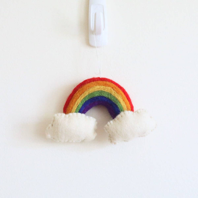 A felt rainbow Christmas ornament hangs from a hook against a white wall. This brightly colored ornament features distinct layers of red, orange, yellow, green, and purple felt. Fluffy white felt clouds are attached to each end, highlighting its three-dimensional design and soft, needle-felted texture for a cheerful touch.