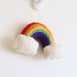 A vibrant handmade felt rainbow Christmas ornament hangs cheerfully from a white hook against a plain white wall. This 3D wool felt ornament features distinct bands of red, orange, yellow, green, blue, and purple. Fluffy white clouds anchor each end of the rainbow, adding to its three-dimensional and whimsical design. It is suspended by a thin, clear string.