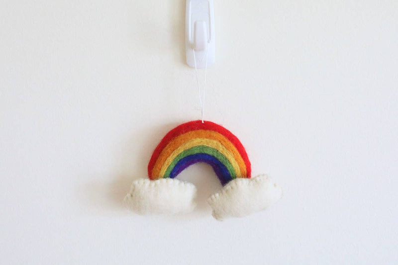A vibrant organic wool felt rainbow Christmas ornament is suspended from a white hook against a bright white wall. This 3D needle-felted ornament features a hand-stitched rainbow arch with distinct red, orange, yellow, green, blue, and purple stripes. Fluffy white clouds are attached at both ends, giving it a soft, textured feel.
