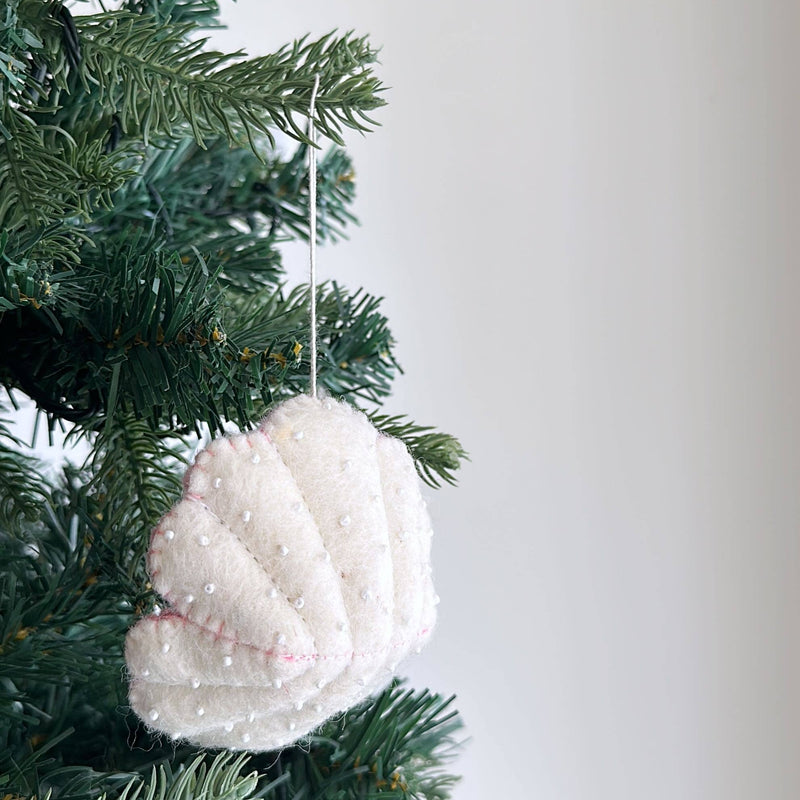 A white handmade wool felt scallop ornament hangs gracefully from a festive Christmas tree branch. This unique felt ornament features delicate rows of tiny white beads along its scalloped edges, subtly outlined with fine pink stitching. A white cotton string secures the charming ornament to the green pine needles, enhancing its rustic elegance.