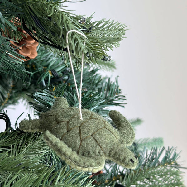 A textured light green wool felt sea turtle ornament hangs gracefully on a Christmas tree. Darker green stitching outlines its shell segments, and a white cotton string is attached for easy hanging, adding rustic charm to the holiday decor.