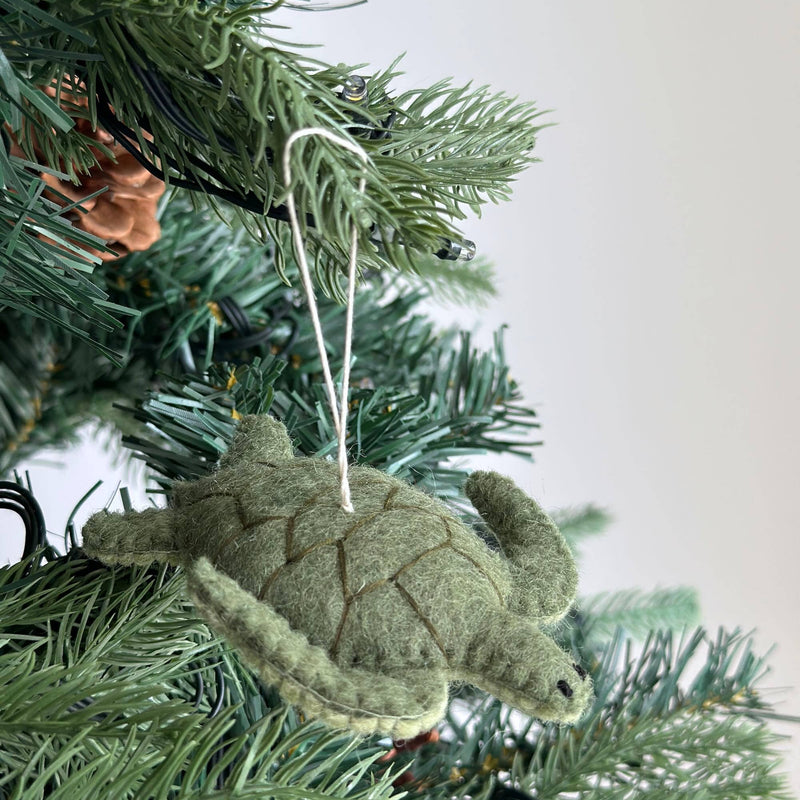 A textured light green wool felt sea turtle ornament hangs gracefully on a Christmas tree. Darker green stitching outlines its shell segments, and a white cotton string is attached for easy hanging, adding rustic charm to the holiday decor.