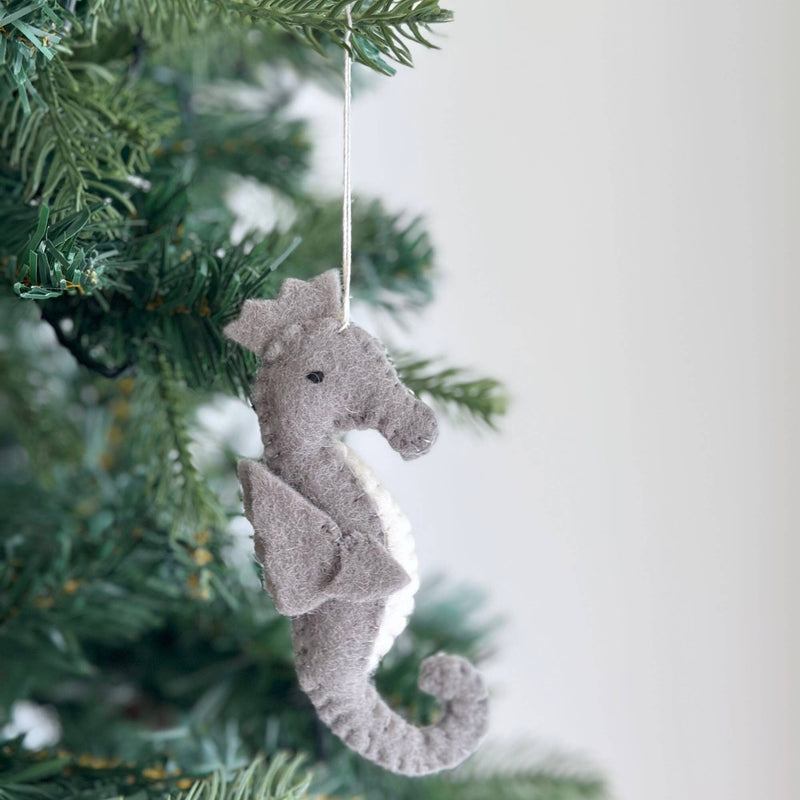 A gray and white wool felt seahorse ornament hangs gracefully on a lush green Christmas tree, set against a plain white wall, creating a heartwarming holiday atmosphere. This handcrafted felt ornament features a textured wool surface, a distinctive curled tail, and a small black eye, suspended by a thin cotton string.