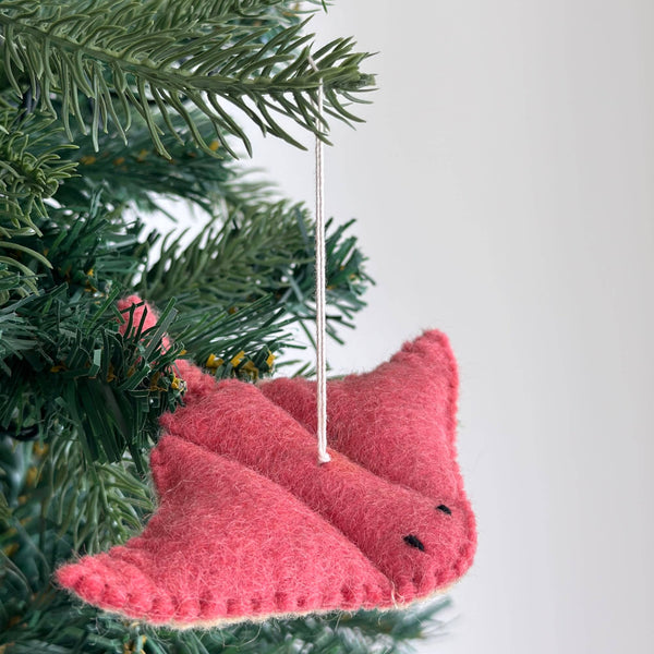 A pink wool felt stingray ornament hangs on a lush green Christmas tree, displayed against a bright white background. This charming sea animal felt ornament features a textured surface, meticulously stitched edges, small black stitched eyes, and a white cotton string for hanging.