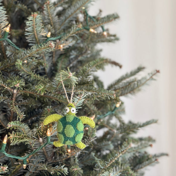 A green felt turtle ornament hangs on a cheerful Christmas tree, sparkling with tiny warm lights. The 3D wool felt turtle features a patterned shell with darker green spots and light green extremities. A jute string is attached for hanging.