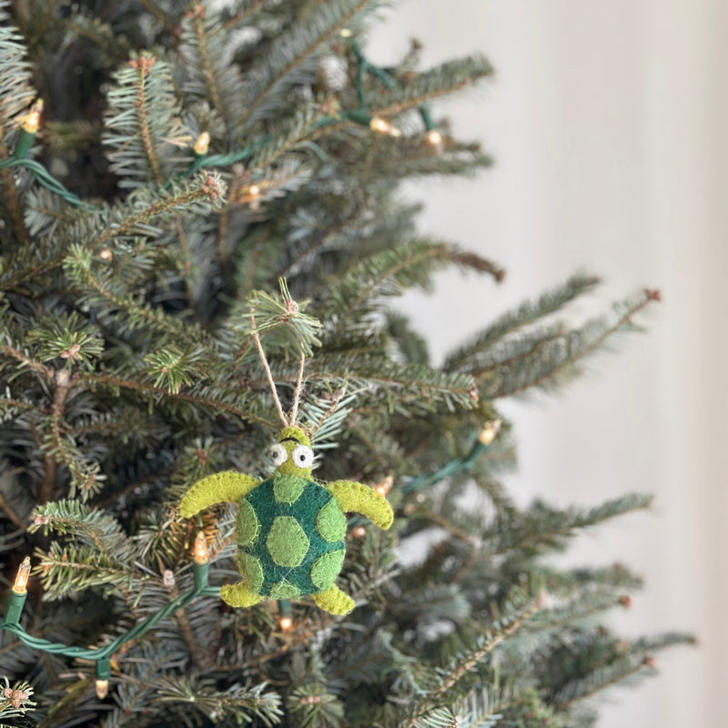 A green felt turtle ornament hangs on a cheerful Christmas tree, sparkling with tiny warm lights. The 3D wool felt turtle features a patterned shell with darker green spots and light green extremities. A jute string is attached for hanging.