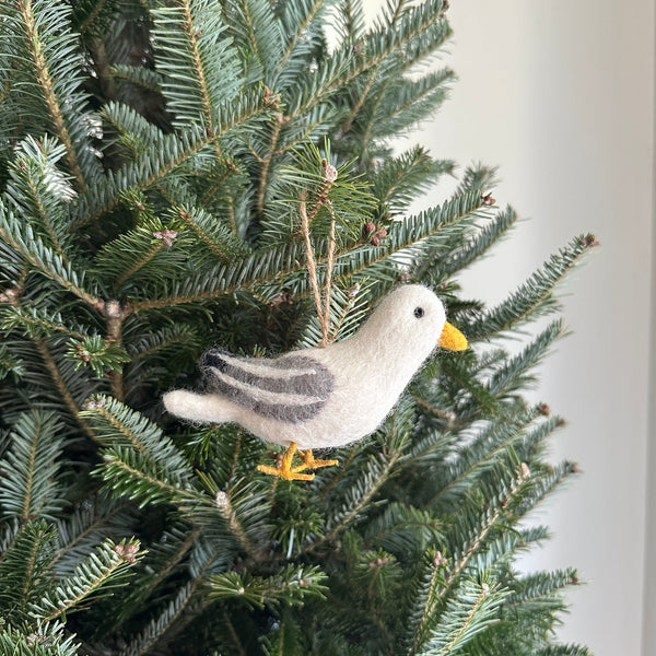A handmade felt seagull ornament hangs from a jute string on a live Christmas tree, set against a blurred light wall. This charming felt seagull features a white body, a yellow beak, and distinctive dark grey markings on its wings. Its textured finish highlights its handcrafted quality, evoking a calm, coastal-inspired Christmas theme.
