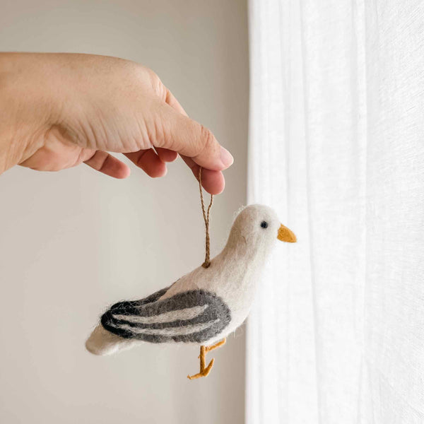 A handmade felt seagull ornament is delicately held by a female hand, suspended by a jute string against a bright, airy background. The felt seagull ornament features a white body with subtle gray stripes on its wings and a yellow beak, highlighting its charming handcrafted feel.