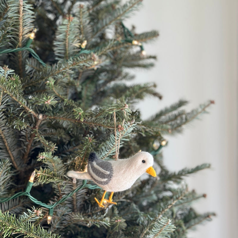 A fluffy felt seagull ornament hangs on a Christmas tree adorned with warm lights. The charming seagull has a pale body with grey wings and tail accents, complemented by a bright yellow beak and feet. A natural jute string is attached for hanging, creating a cozy and festive holiday scene.