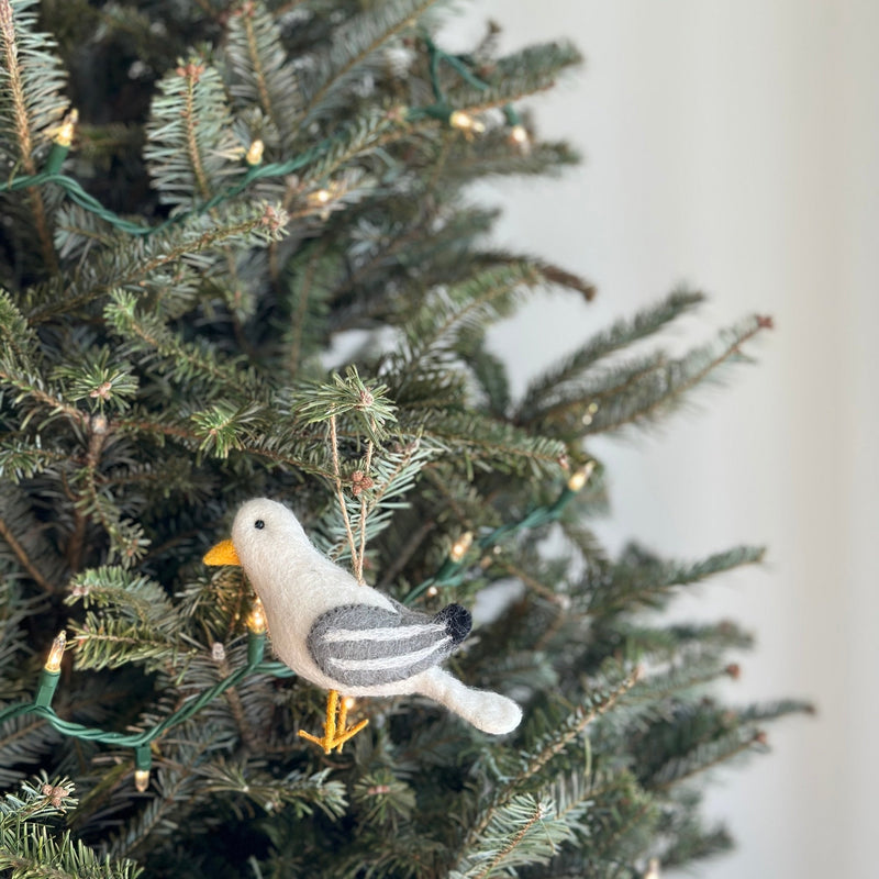 A felt seagull ornament hangs from a lit Christmas tree branch. The unique Christmas ornament features a fluffy white body, gray wings with darker gray stripes, black eyes, and a yellow beak and feet. A jute string allows the handcrafted felt ornament to gracefully hang amidst the evergreen needles and soft lights.