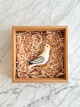 A handcrafted seagull felt ornament is nestled in a brown cardboard box, filled with light-brown paper, against a bright marble backdrop. This charming felt seagull ornament features a white body with grey and white striped wings and tail, a yellow beak and feet, and black eyes. A jute string on its head allows for hanging.