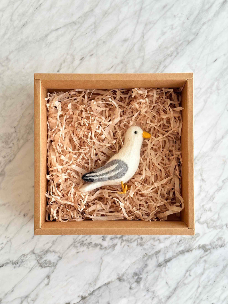 A handcrafted seagull felt ornament is nestled in a brown cardboard box, filled with light-brown paper, against a bright marble backdrop. This charming felt seagull ornament features a white body with grey and white striped wings and tail, a yellow beak and feet, and black eyes. A jute string on its head allows for hanging.