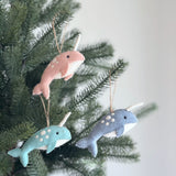 Three colorful felt narwhal ornaments hang from Christmas tree branches with rustic twine, creating a whimsical, festive atmosphere. Each unique narwhal ornament features white spots, a spiraled horn, and a white underbelly that contrasts with its vibrant upper hue of baby pink, ocean blue, or light baby blue, completed by small dark eyes.