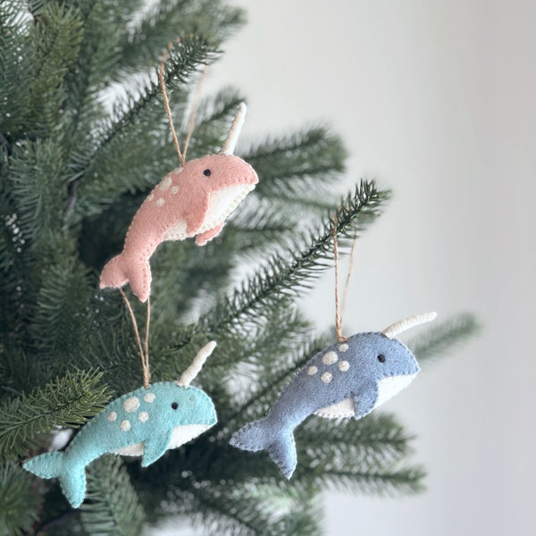 Three colorful felt narwhal ornaments hang from Christmas tree branches with rustic twine, creating a whimsical, festive atmosphere. Each unique narwhal ornament features white spots, a spiraled horn, and a white underbelly that contrasts with its vibrant upper hue of baby pink, ocean blue, or light baby blue, completed by small dark eyes.