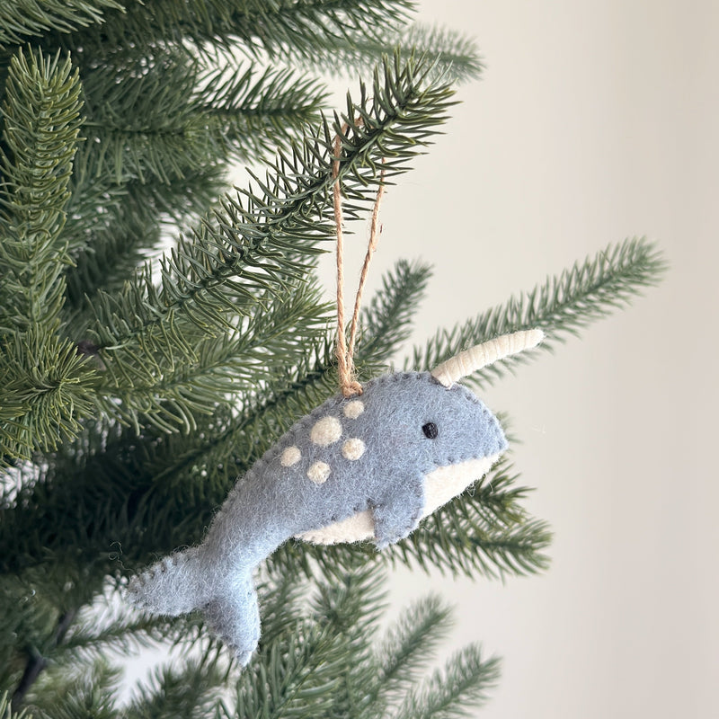 A light blue felt narwhal ornament hangs festively on a Christmas tree. This charming felted narwhal features white spots, a white horn, and a white belly, suspended by a brown Jute string.