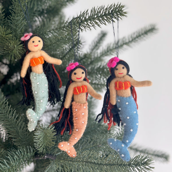 Three vibrant Silk Sea Mermaid felt ornaments are shown hanging joyfully on a green Christmas tree branch. Each handmade felt ornament features flowing black yarn hair, stitched orange and yellow seashell tops, and tails in light blue, peach, and light green adorned with white pearls. A clear string attaches them to the branch, set against a soft white background.