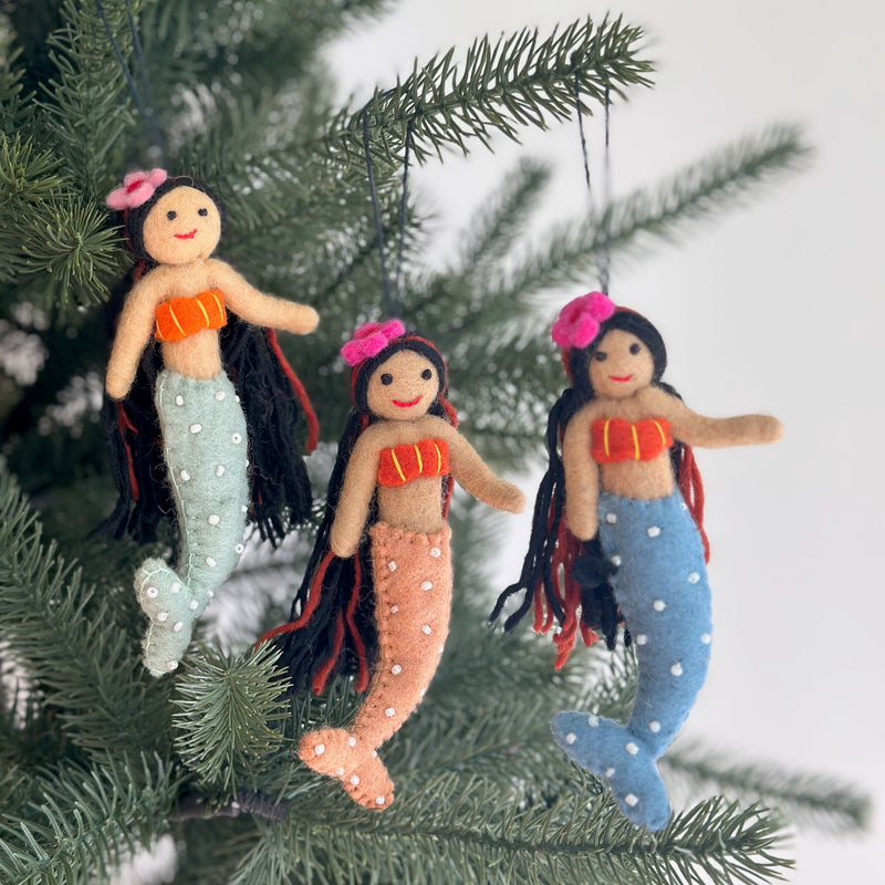 Three vibrant Silk Sea Mermaid felt ornaments are shown hanging joyfully on a green Christmas tree branch. Each handmade felt ornament features flowing black yarn hair, stitched orange and yellow seashell tops, and tails in light blue, peach, and light green adorned with white pearls. A clear string attaches them to the branch, set against a soft white background.