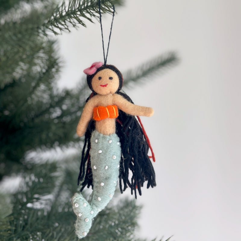 A Silk Sea felt mermaid ornament hangs gracefully on a Christmas tree. This unique handmade ornament features a soft tan face with a stitched red smile and flowing black yarn hair. It has an orange seashell top and a light blue-green tail adorned with white pearl-like embellishments. A black cotton string is attached at the top, ready for display.
