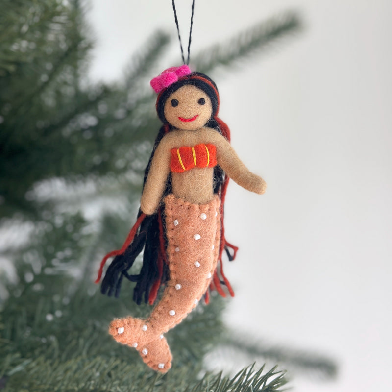 A silk sea mermaid felt ornament is suspended from a green Christmas tree branch against a soft white backdrop. This joyful handmade felt ornament has a tan upper body with an orange seashell top detailed with yellow lines. It features flowing black yarn hair and a peach-colored mermaid tail adorned with small white pearl-like dots.