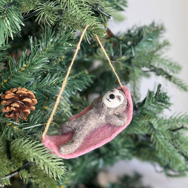 A wool felt sloth ornament hangs on a Christmas tree, its jute string loop suspended from a green branch adorned with a pine cone. The grey sloth lounges comfortably in a pink hammock, featuring a light grey face with a small black stitched smile. Its body is crafted from textured light and dark grey wool felt, enhancing its charming, relaxed appearance.