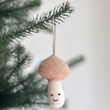 A handcrafted needle felted smiley beige mushroom ornament hangs from a lush green tree branch. This charming mushroom felt ornament features a distinctive beige cap and a creamy white stem adorned with a cheerful smile. A jute string is visible for hanging.